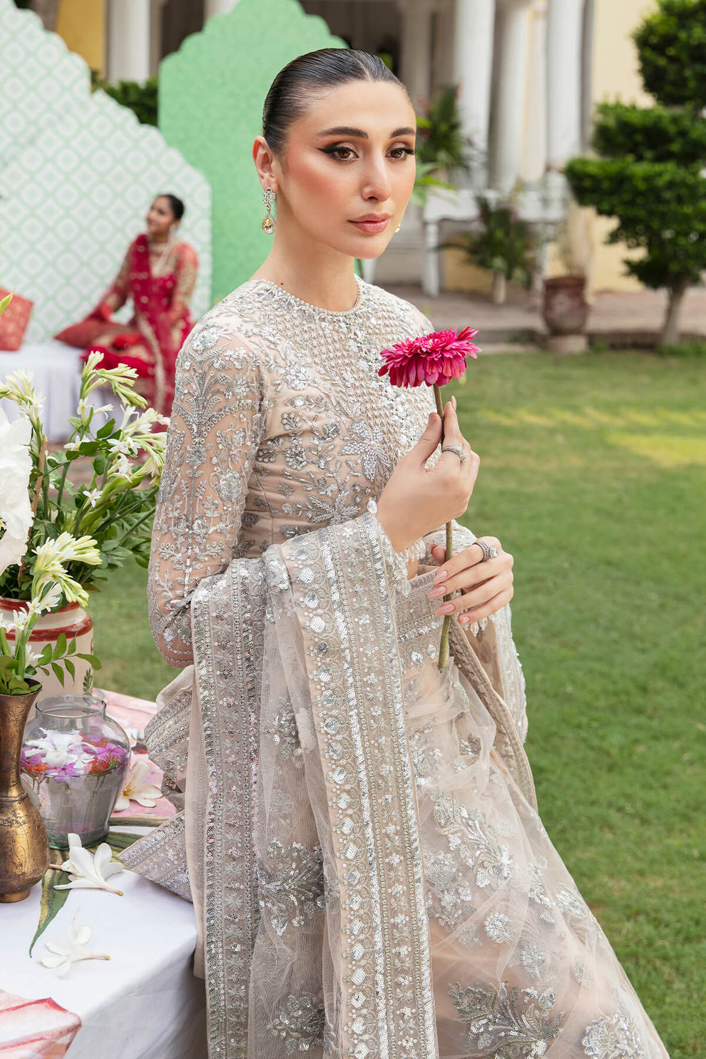 Woman in a silver embroidered outfit holding a pink flower in an outdoor setting.