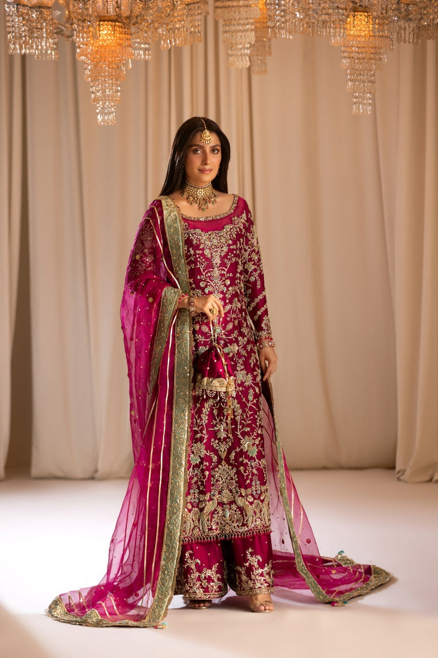 Woman in a traditional pink and gold embroidered outfit with a matching dupatta, standing against a beige curtain.