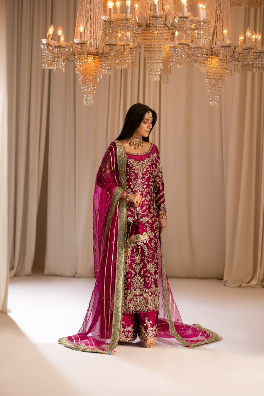 Woman in a traditional pink and red embroidered outfit standing in front of a chandelier.